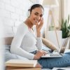 A imagem mostra uma jovem sorridente sentada confortavelmente em uma cama, usando fones de ouvido brancos e um notebook no colo. Ela está em um ambiente iluminado e acolhedor, com decoração suave, incluindo uma planta no fundo e uma parede de tijolos brancos. A cena transmite leveza, concentração e bem-estar, sugerindo um momento de estudo, trabalho remoto ou lazer digital.