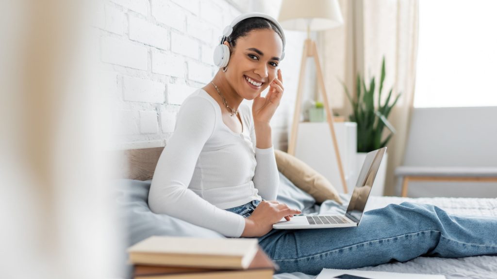 A imagem mostra uma jovem sorridente sentada confortavelmente em uma cama, usando fones de ouvido brancos e um notebook no colo. Ela está em um ambiente iluminado e acolhedor, com decoração suave, incluindo uma planta no fundo e uma parede de tijolos brancos. A cena transmite leveza, concentração e bem-estar, sugerindo um momento de estudo, trabalho remoto ou lazer digital.
