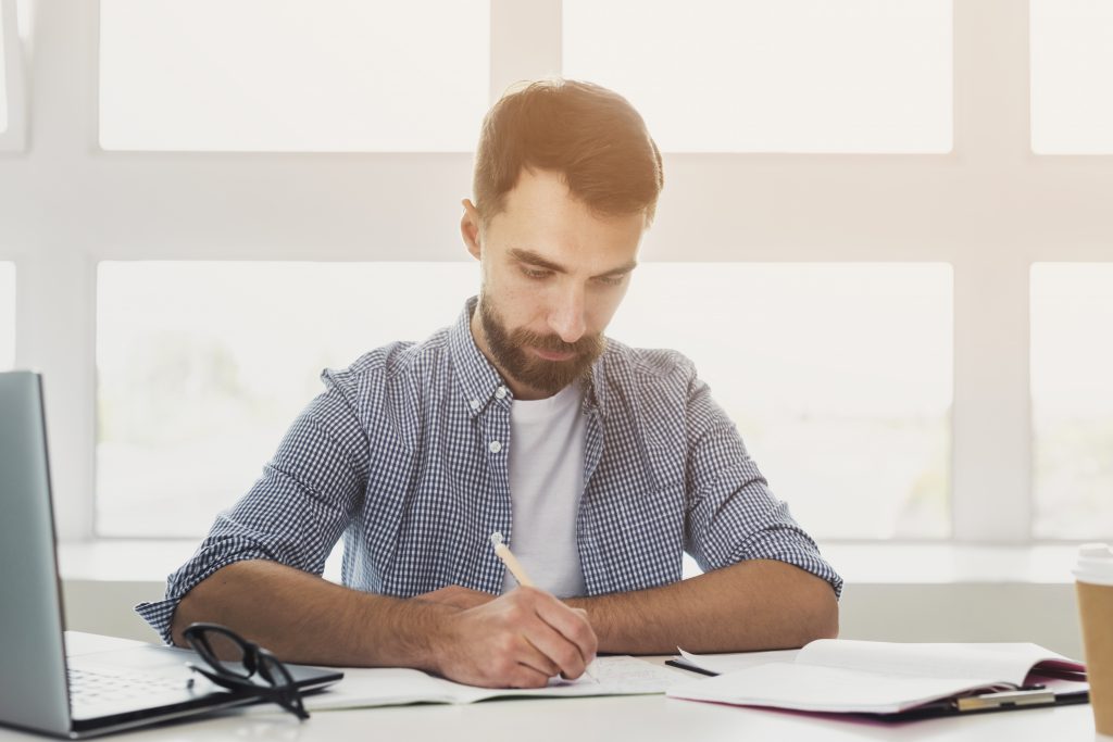 Homem estudando, é possível ver um laptop, cadernos e um óculos de grau na mesa