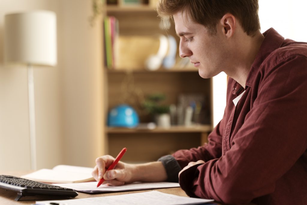A imagem mostra um homem jovem, sentado à mesa, concentrado em seus estudos. Ele está escrevendo com uma caneta vermelha em uma folha de papel enquanto observa atentamente o que está escrevendo. Ao fundo, é possível ver uma estante de livros e outros itens organizados, criando um ambiente de estudo tranquilo e focado. Ele usa uma camisa vermelha, e a luz suave que entra pela janela ilumina a cena, dando uma sensação de aconchego e concentração.