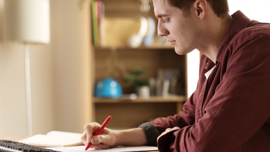 A imagem mostra um homem jovem, sentado à mesa, concentrado em seus estudos. Ele está escrevendo com uma caneta vermelha em uma folha de papel enquanto observa atentamente o que está escrevendo. Ao fundo, é possível ver uma estante de livros e outros itens organizados, criando um ambiente de estudo tranquilo e focado. Ele usa uma camisa vermelha, e a luz suave que entra pela janela ilumina a cena, dando uma sensação de aconchego e concentração.