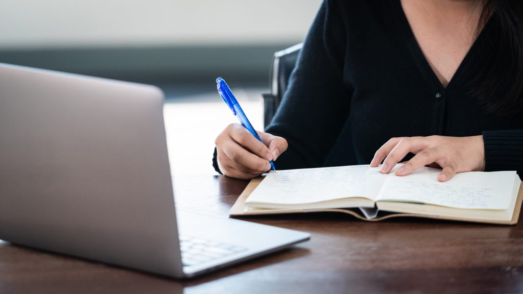 A imagem mostra uma pessoa escrevendo em um caderno com uma caneta azul, enquanto consulta algo em um notebook aberto à sua frente. A pessoa veste um suéter preto e está focada na tarefa, indicando que está respondendo questões ou fazendo anotações, possivelmente em um ambiente de estudo ou trabalho. A cena transmite concentração e organização.
