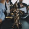 A imagem mostra uma estátua da Justiça, representada por uma mulher com os olhos vendados e segurando uma balança de equilíbrio, simbolizando a imparcialidade. A estátua está em primeiro plano, com advogados em traje formal ao fundo, em um ambiente de escritório, onde documentos e ferramentas de trabalho estão visíveis. O foco é na balança, destacando o tema da justiça e da responsabilidade legal.