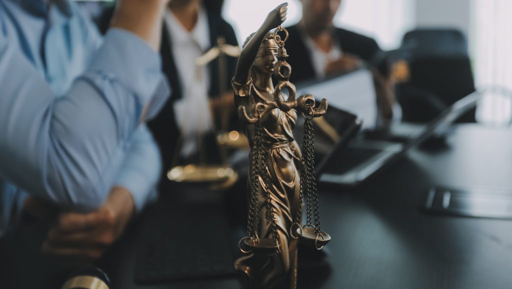 A imagem mostra uma estátua da Justiça, representada por uma mulher com os olhos vendados e segurando uma balança de equilíbrio, simbolizando a imparcialidade. A estátua está em primeiro plano, com advogados em traje formal ao fundo, em um ambiente de escritório, onde documentos e ferramentas de trabalho estão visíveis. O foco é na balança, destacando o tema da justiça e da responsabilidade legal.