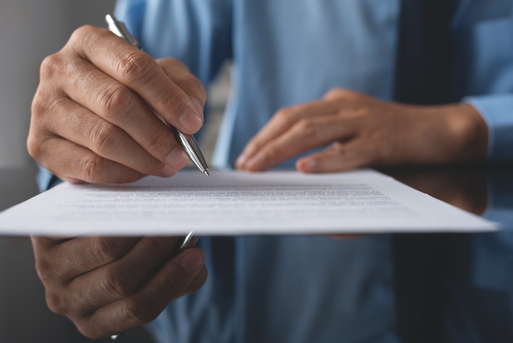 A imagem mostra um profissional com uma camisa azul, assinando um documento com uma caneta prata. O papel está posicionado sobre uma superfície reflexiva, o que cria um reflexo visível na parte inferior do documento. O ambiente parece ser formal, focado em um momento de revisão ou assinatura de documentos importantes.