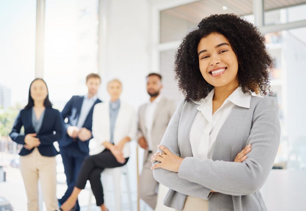 A imagem mostra uma mulher sorridente em primeiro plano, vestindo um terno cinza e com os braços cruzados, enquanto posa em um ambiente de escritório moderno. Ela está à frente de um grupo de colegas de trabalho, que aparecem desfocados ao fundo. Todos estão vestidos de forma profissional e o ambiente é bem iluminado, com janelas grandes que proporcionam luz natural. A mulher transmite confiança e postura profissional.