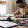 A imagem mostra um homem sentado à mesa, concentrado em seus estudos. Ele usa fones de ouvido e está escrevendo em um caderno enquanto observa seu computador portátil aberto. O ambiente é iluminado, com luz natural entrando pela janela ao fundo, criando uma atmosfera tranquila e produtiva. Há vários materiais de estudo espalhados pela mesa, como cadernos, post-its coloridos e outros itens, refletindo um ambiente organizado de trabalho.