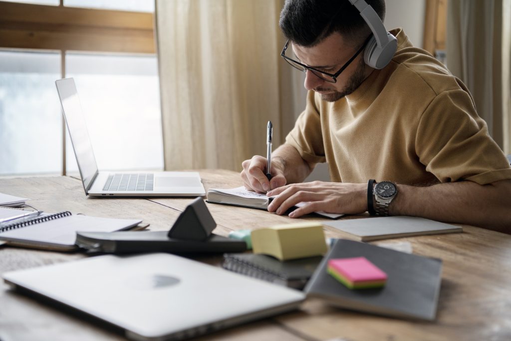 A imagem mostra um homem sentado à mesa, concentrado em seus estudos. Ele usa fones de ouvido e está escrevendo em um caderno enquanto observa seu computador portátil aberto. O ambiente é iluminado, com luz natural entrando pela janela ao fundo, criando uma atmosfera tranquila e produtiva. Há vários materiais de estudo espalhados pela mesa, como cadernos, post-its coloridos e outros itens, refletindo um ambiente organizado de trabalho.