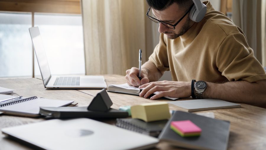 A imagem mostra um homem sentado à mesa, concentrado em seus estudos. Ele usa fones de ouvido e está escrevendo em um caderno enquanto observa seu computador portátil aberto. O ambiente é iluminado, com luz natural entrando pela janela ao fundo, criando uma atmosfera tranquila e produtiva. Há vários materiais de estudo espalhados pela mesa, como cadernos, post-its coloridos e outros itens, refletindo um ambiente organizado de trabalho.