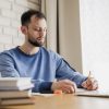 Homem sentado a mesa fazendo anotações em um cadern. É possível ver uma pilha de livros no canto da imagem