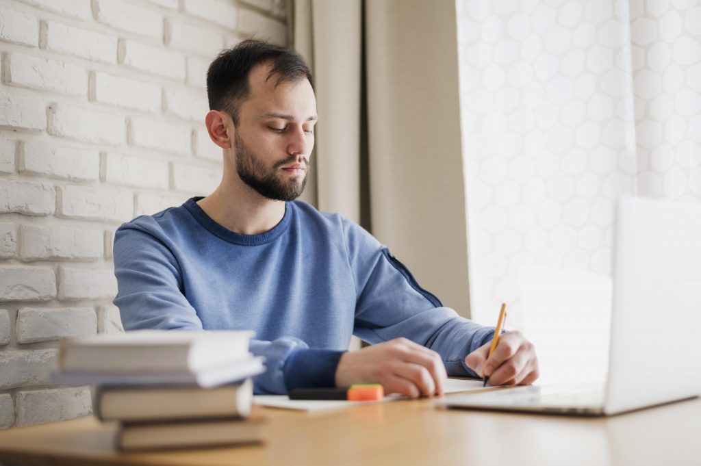 Homem sentado a mesa fazendo anotações em um cadern. É possível ver uma pilha de livros no canto da imagem