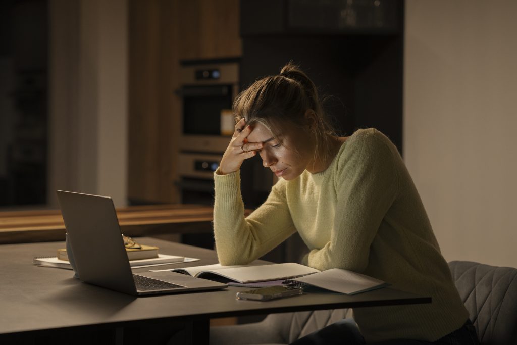 Mulher com expressão cansada estuda à noite em uma cozinha, com a mão na testa, cercada por cadernos e notebook sobre a mesa.