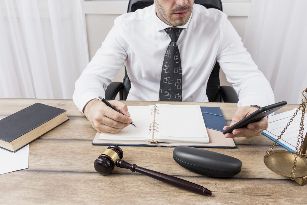 Homem de terno trabalhando em escritório jurídico, fazendo anotações em caderno enquanto consulta o celular, com martelo do juiz e balança da justiça sobre a mesa.