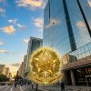 Fachada imponente do prédio do Tribunal Regional Federal da 3ª Região (TRF3), um arranha-céu espelhado em uma avenida movimentada. Centralizado na imagem, o Brasão da República Federativa do Brasil aparece em destaque dourado sobreposto à arquitetura.