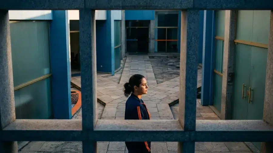 Mulher em perfil atravessando pátio de prédio público com caminhos de pedra e luz lateral